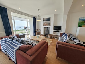 A cozy living room with a sofa, armchair, and a television. There's a fireplace, a telescope, and a view of the sea through large windows. - 3 Bed in Craster (oc-c25115) (Dunstan)