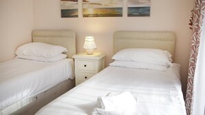 A hotel room with two beds, a nightstand with a lamp, and a painting of a sunset over the ocean. - 2 Bed in Mumbles (oc-hh120) (The Mumbles)