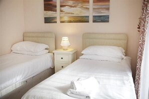 A hotel room with two beds, a nightstand with a lamp, and a painting of a sunset over the ocean. - 2 Bed in Mumbles (oc-hh120) (The Mumbles)