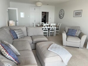 A living room with a sectional sofa, a dining table with chairs, a clock on the wall, and a door leading to another room.