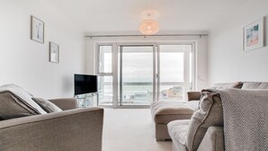 Ett vardagsrum med en tv, två soffor, ett soffbord i glas och utsikt över havet genom stora glasdörrar.