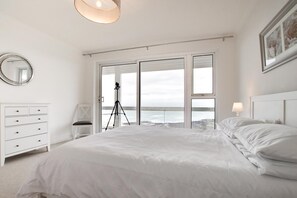 A bedroom with a large bed, a tripod, a chair, and a view of the sea through large windows.