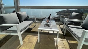 A balcony with a view of the sea, a table with a bottle and glasses, and two white chairs.