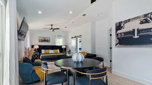 A modern living room with a round black table and matching chairs, a sofa with yellow cushions, and a large painting of a car on the wall. - The Augustine Suite l Heated Pool  l Walk to DNTN (St. Augustine)