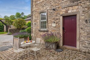 Ein Steingebäude mit einer roten Tür, einem Fenster mit Fensterläden und einem kleinen Garten mit violetten Blumen.