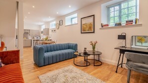 Een moderne woonkamer met een blauwe bank, een kleine ronde tafel en een grijze fauteuil. Achtergrond is een keuken met witte kasten en een eethoek.