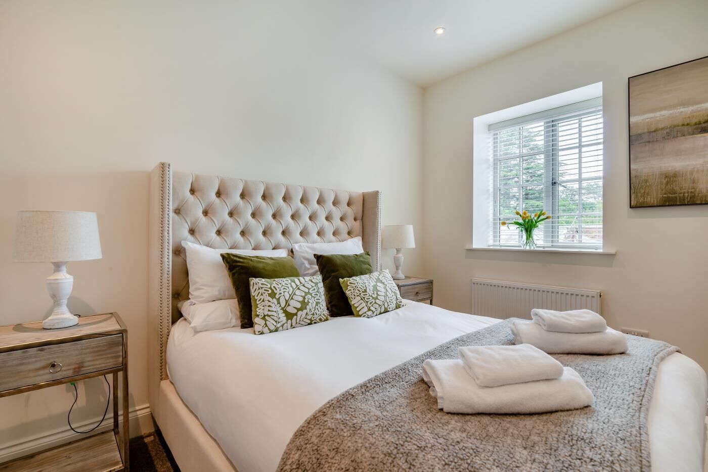 A neatly made bed with a tufted headboard, a bedside table with a lamp, and a window with a view of greenery.
