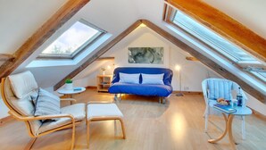 Un dormitorio estilo loft acogedor con techo inclinado, un sofá azul, dos sillones, una mesita y un ventanal grande.