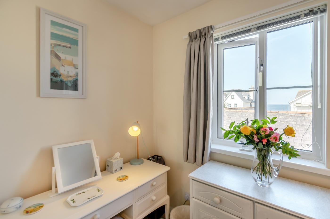 A bedroom with a desk, a bedside lamp, a framed picture, a vase with flowers, and a window with curtains.