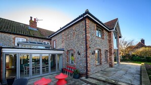 A stone building with a patio area featuring red tables and chairs, a glass-enclosed dining area, and a stone pathway leading to the entrance. - 4 Bed in Blakeney (oc-2070) (Blakeney)