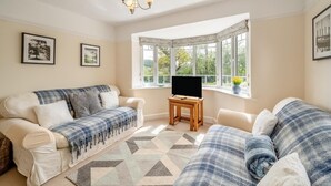 A cozy living room with a sofa set, a television on a wooden stand, and a bay window with a view of greenery.