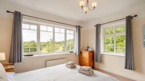 A bedroom with a large bed, two windows with curtains, a wooden dresser, and a chandelier.