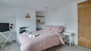 A bedroom with a bed, a TV on a stand, a shelf with a picture, and a wooden door. - 2 Bed in Aldeburgh (oc-3al) (Aldeburgh)