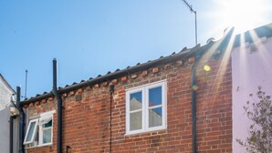 A two-story brick house with a green door, a small patio with a table and chairs, and a pink wall on the right. - 2 Bed in Aldeburgh (oc-3al) (Aldeburgh)