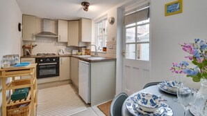 A compact kitchen with a stove, oven, and sink, a dining area with a table set for two, and a window with curtains. - 2 Bed in Aldeburgh (oc-3al) (Aldeburgh)