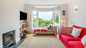 Een woonkamer met een open haard, een rode bank, een televisie en een groot raam met uitzicht op groen.