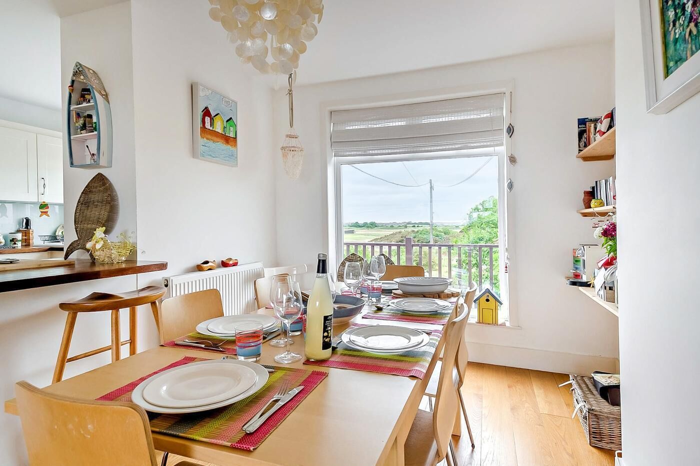 A dining area with a table set for four, a kitchen in the background, a view through a window, and a small yellow birdhouse on the windowsill.