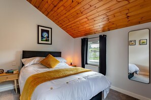 Une chambre avec un plafond en bois, un lit avec une literie blanche et un oreiller jaune, une table de chevet avec des livres, une commode, une fenêtre avec des rideaux et un miroir sur le mur.