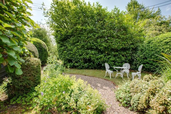 A garden with a gravel path, white metal chairs, and a hedge.