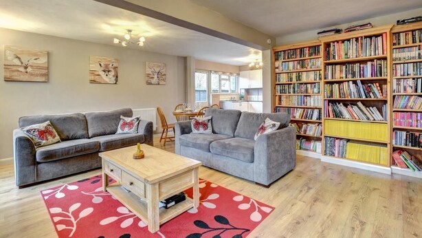 Ein geräumiges Wohnzimmer mit zwei grauen Sofas, einem Holztisch und einem roten Teppich mit floralem Muster. Eine große Bücherwand voller Bücher befindet sich rechts.