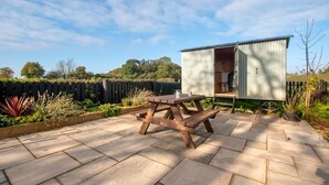 Eine Terrasse mit Holztisch und Bänken, ein kleines Gartenhaus und ein Garten mit einer Umzäunung.