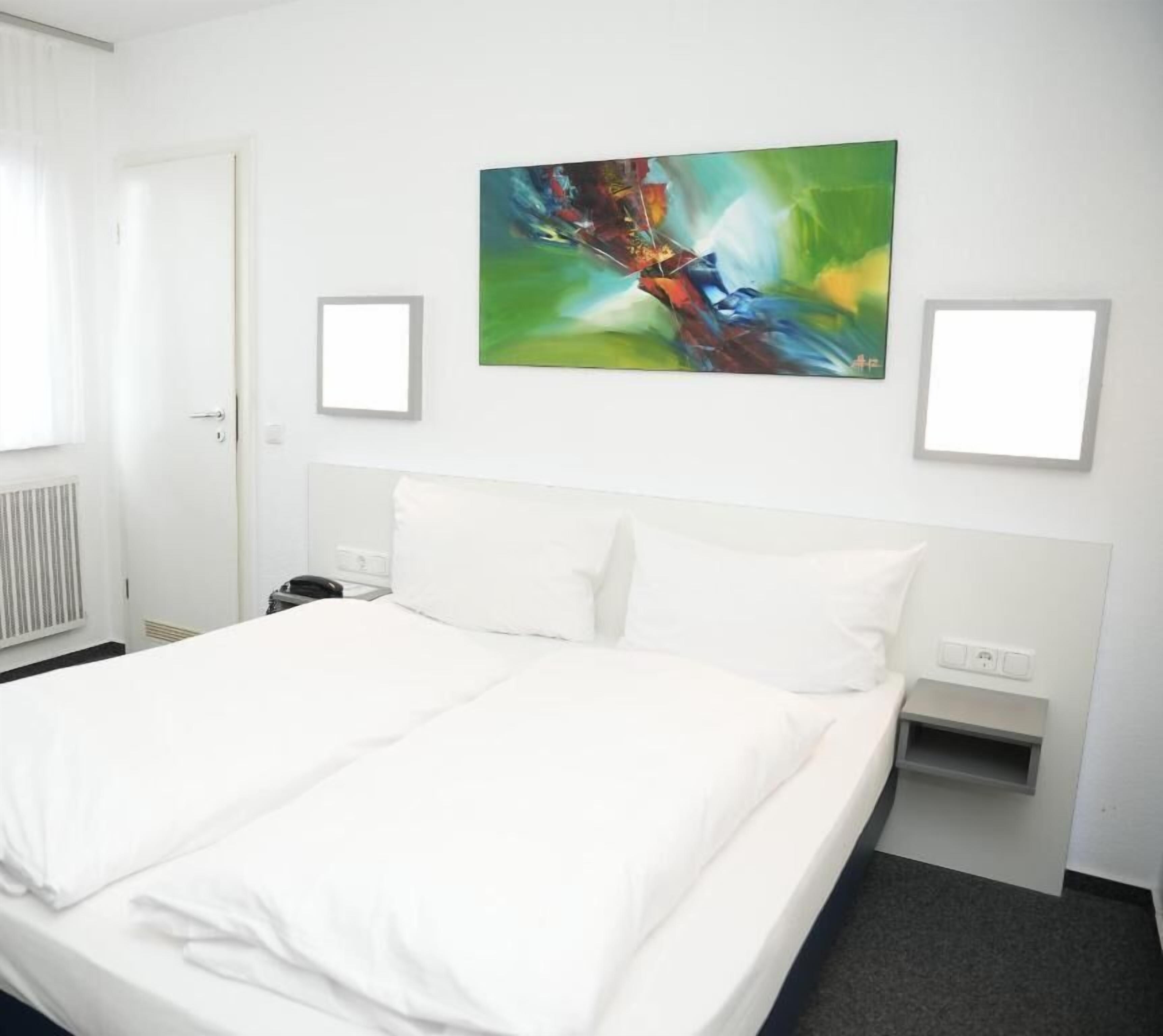 A hotel room with a large bed, a nightstand, a wall-mounted painting, and a window with curtains.