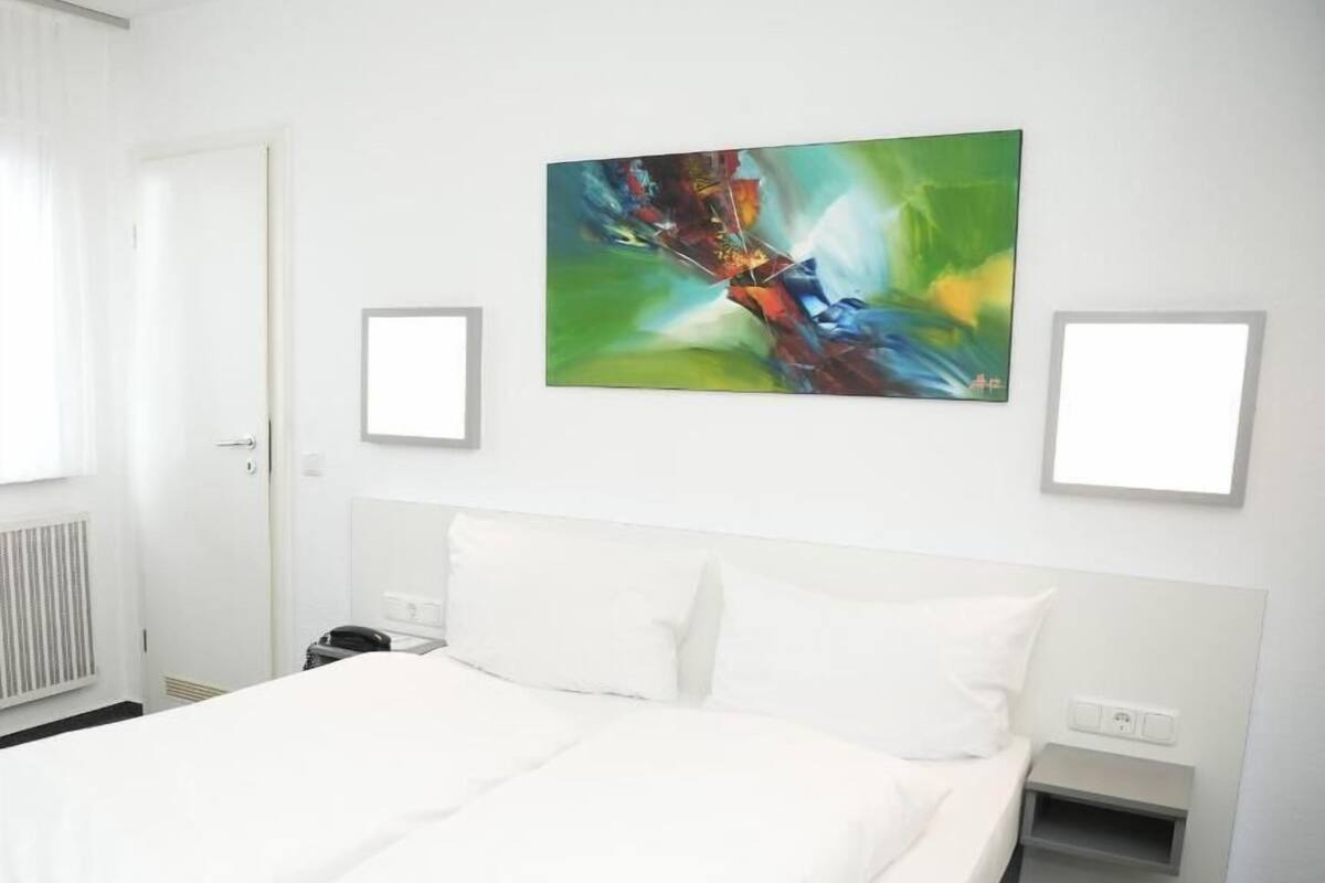 A hotel room with a large bed, a nightstand, a wall-mounted painting, and a window with curtains.