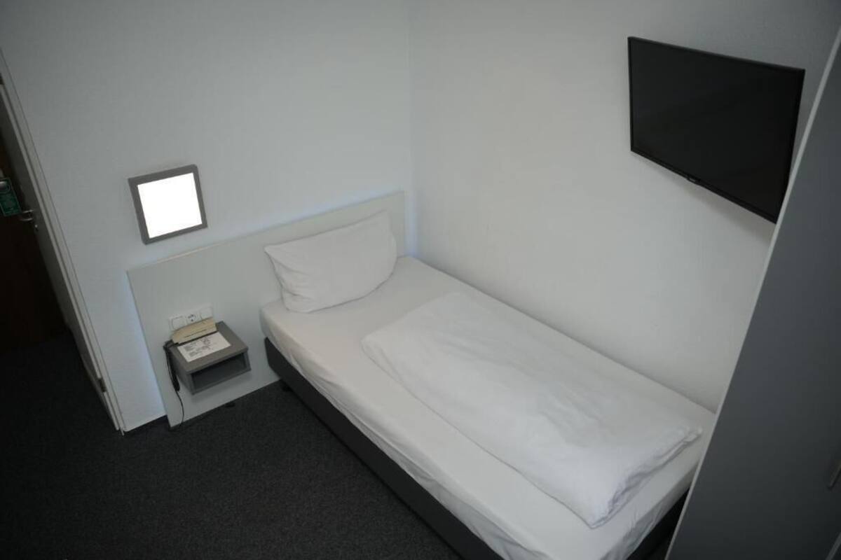 A small, single-bed room with a white bed, a nightstand, a bedside lamp, and a flat-screen TV mounted on the wall.