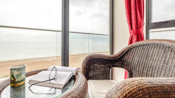 A room with a large window overlooking a beach, a wicker chair, a glass table with a book and glasses, and a red curtain.