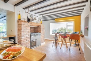 Eine Küche mit Ziegelofen, Holzhockern und einem Esstisch mit Blick ins Freie.