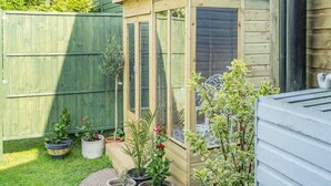Un cobertizo de madera con una ventana, rodeado de plantas en macetas y una valla verde.