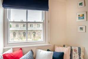 Ein Zimmer mit großem Fenster mit Blick auf ein historisches Gebäude, einer gepolsterten Bank mit Kissen und drei gerahmten Bildern an der Wand.