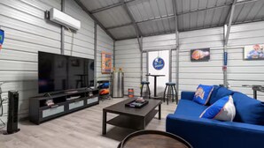 A spacious industrial-style room with a large flat-screen TV, a blue sofa with cushions, a coffee table, and a small bar area with stools. - The Gator Den: Perfect for Groups & Families! 3 BR/ 2 Bath (Gainesville)