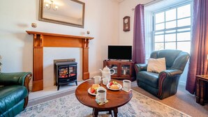 Ein gemütliches Wohnzimmer mit Kamin, Fernseher, Ledersessel und Esstisch mit Kaffeekanne und Gebäck.