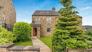 A stone house with a paved pathway leading to the entrance, surrounded by well-maintained hedges and a tall coniferous tree. - 3 Bed in Hawes (oc-ds967) (Hawes)