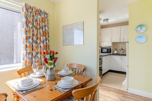 A dining area with a table set for four, a vase of flowers, and a kitchen with white cabinets and a microwave.