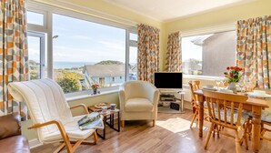 Ein Zimmer mit Fernseher, einem Esstisch mit Stühlen, einer Couch und großen Fenstern mit Vorhängen.