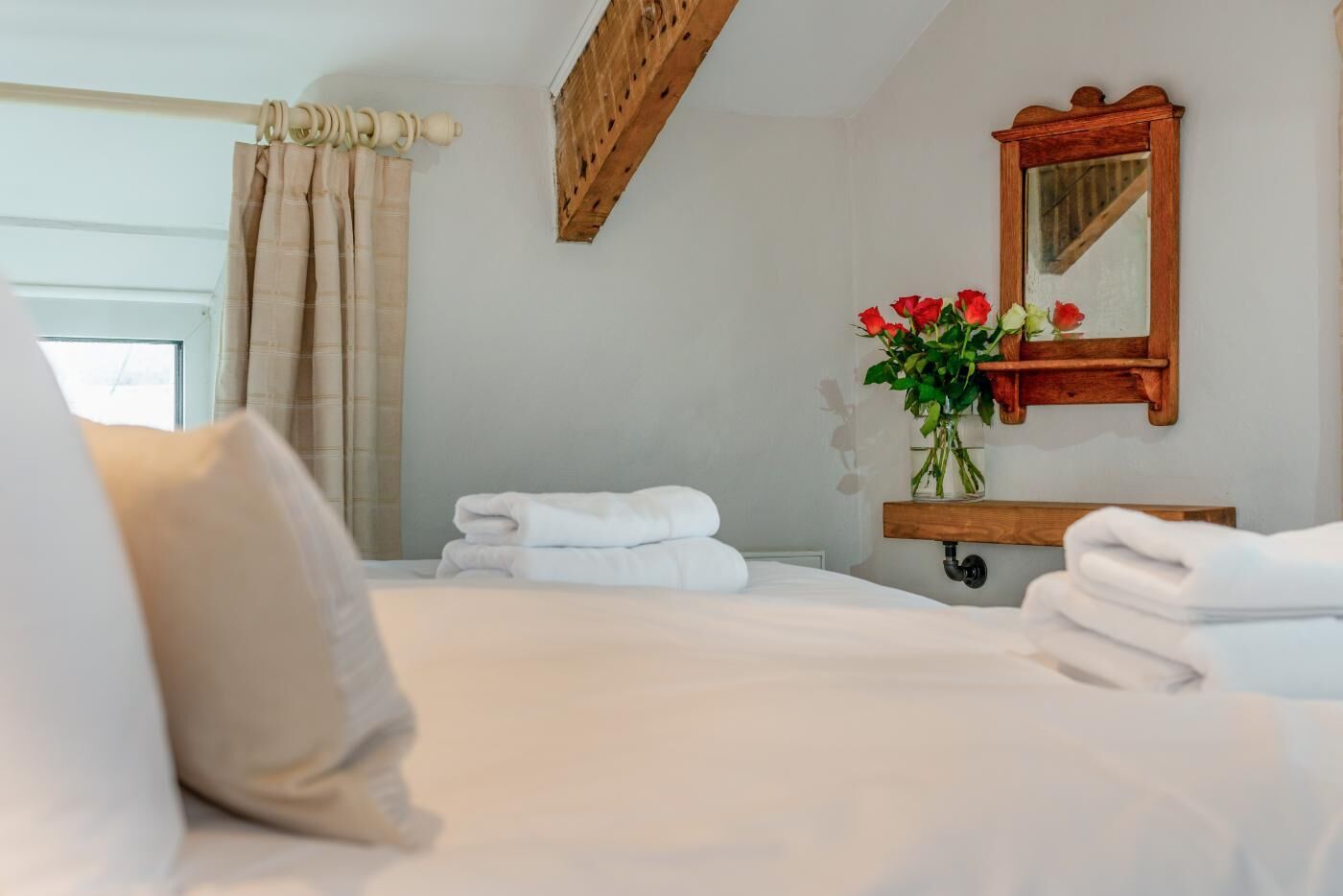 Une chambre d’hôtel avec un lit, une étagère en bois avec des serviettes, un miroir et un vase avec des fleurs.