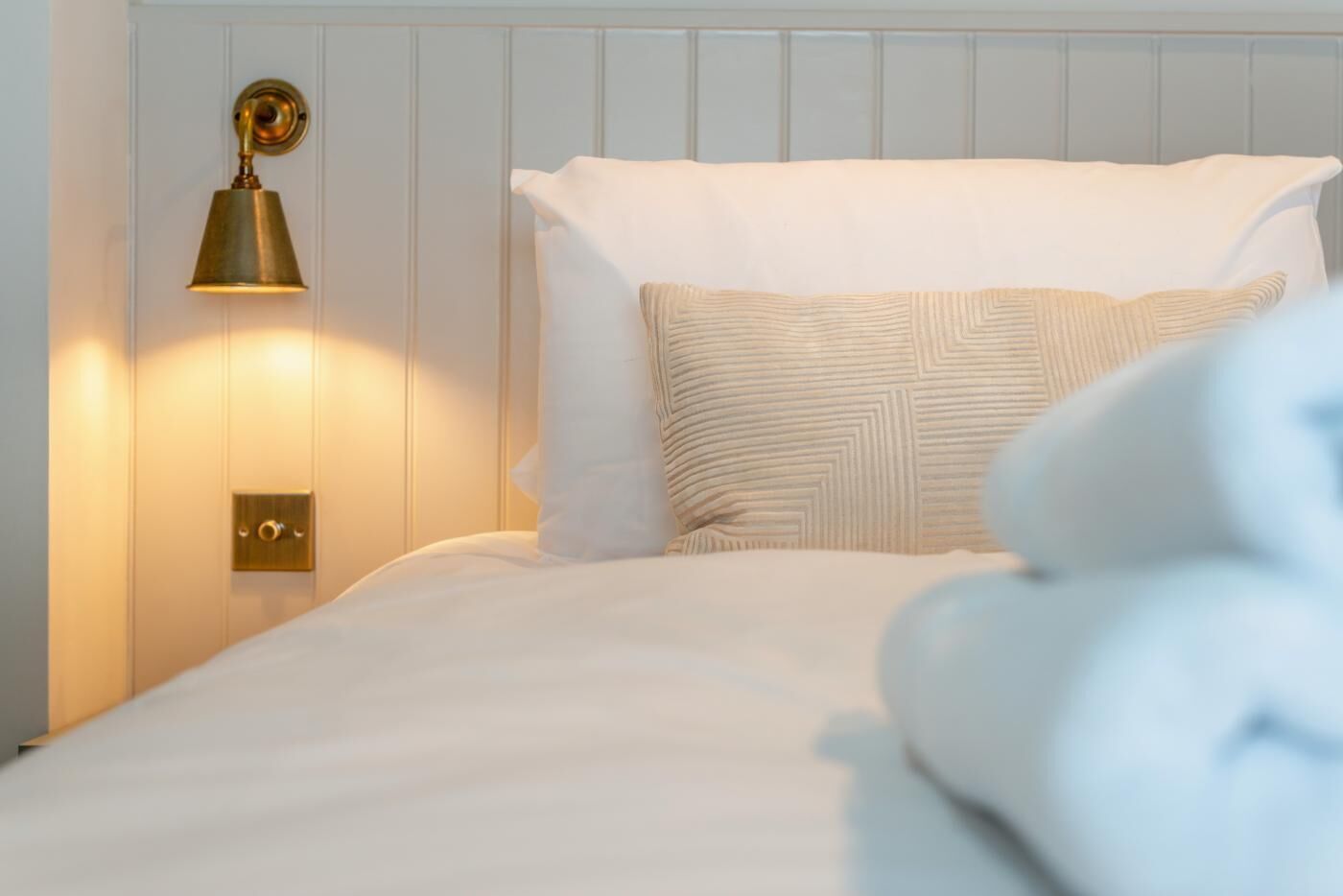 Una cama bien hecha con sábanas blancas y un cojín estampado, una lámpara de pared de latón y una puerta con un pomo sencillo.