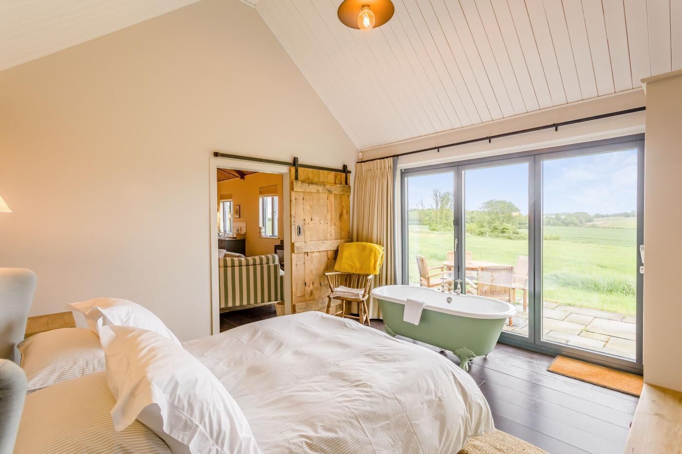 A bedroom with a bed, a chair, a bathtub, and a sliding door leading to a balcony with a view of the countryside.