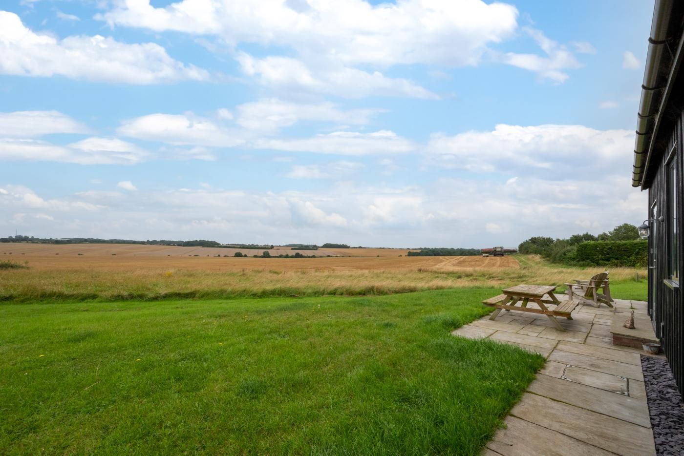 Udsigt over en græsmark med et picnicbord og stole på en træterrasse, med et stort, åbent landskab foran.
