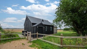 A black wooden building with a gravel path leading to it, surrounded by greenery and a wooden fence. - 1 Bed in Wickhambrook (oc-tosh) (Wickhambrook)