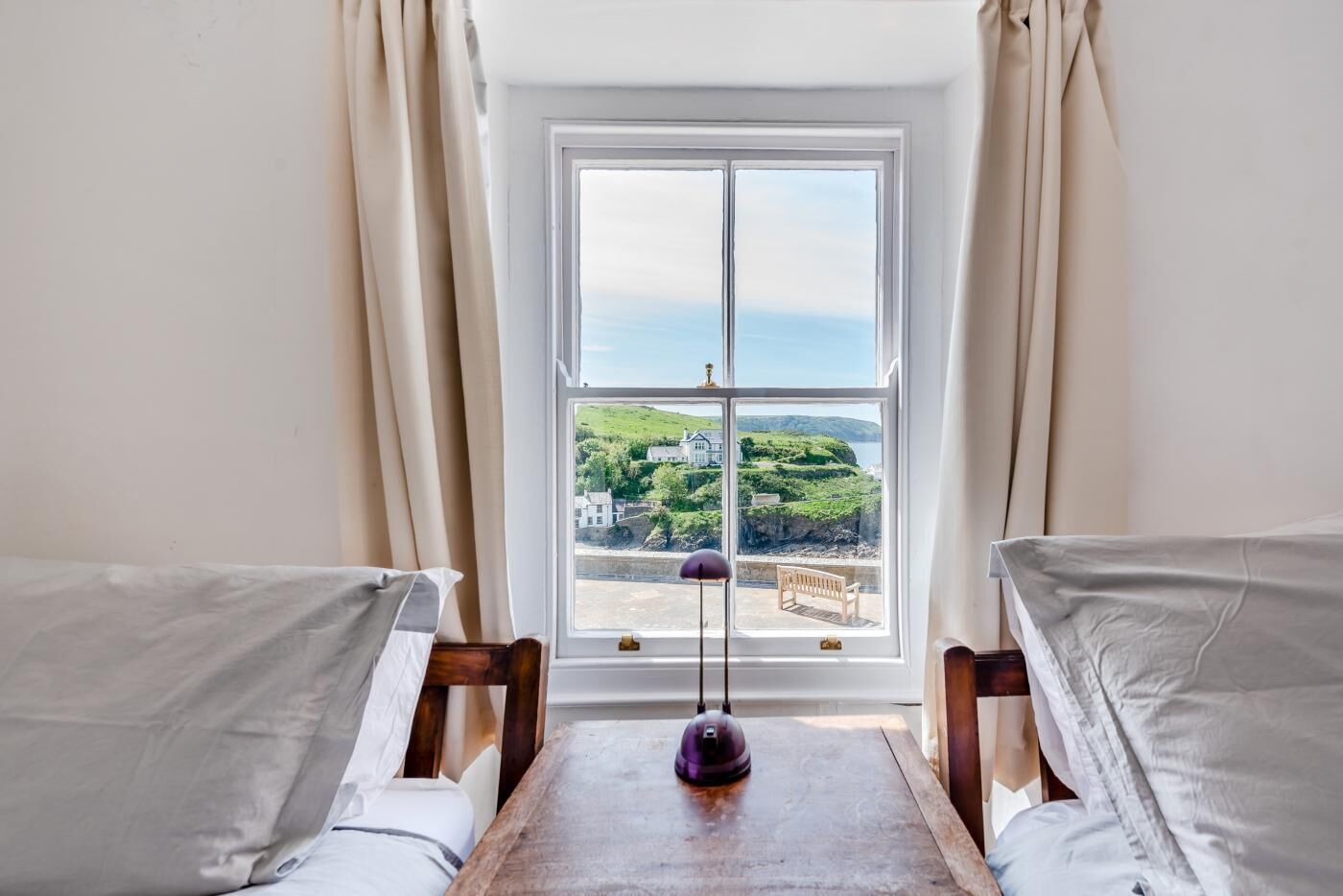 A room with two beds, a wooden table, a lamp, and a window with a view of a green landscape and buildings.