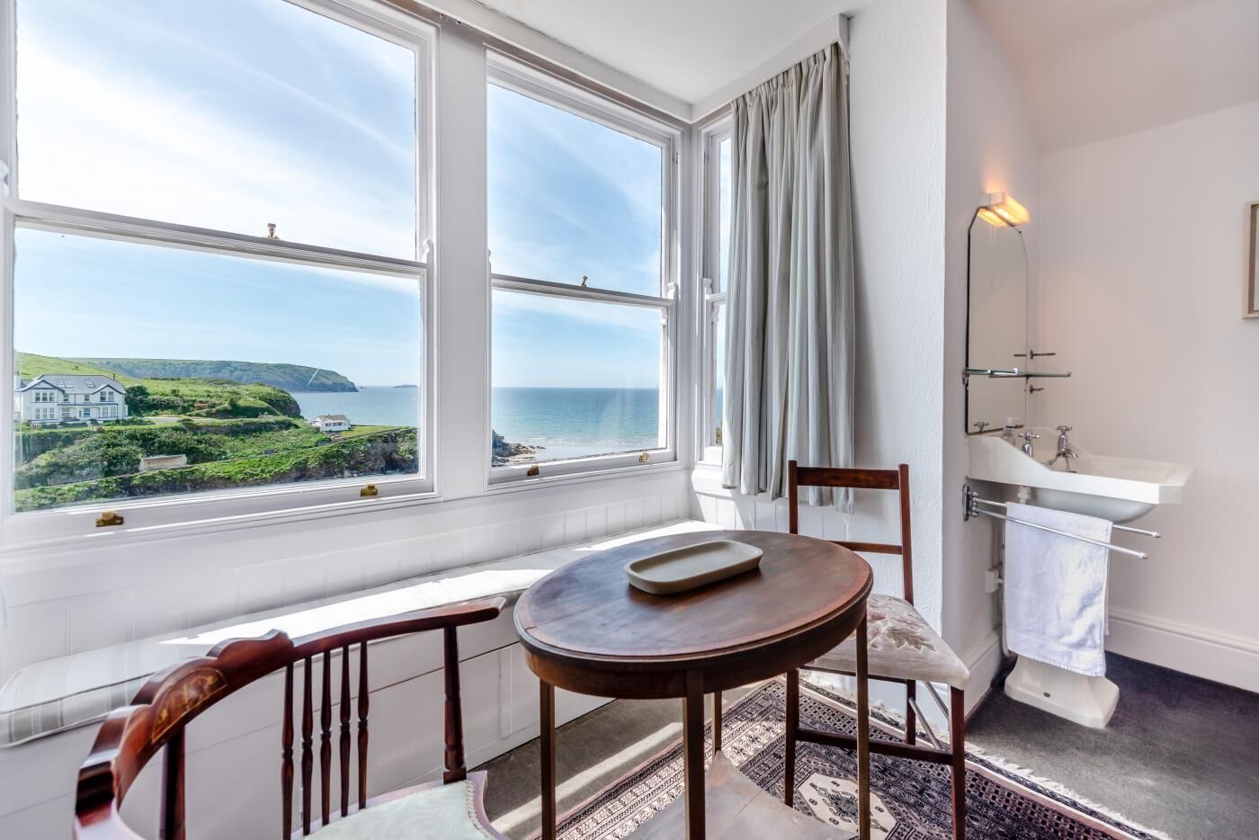 Een kamer met uitzicht op zee, een ronde tafel met dienblad, een stoel, een wastafel en een raam met gordijnen.