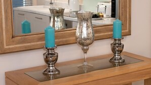 A wooden console table with three teal candles on silver candle holders, a glass vase, and a mirror reflecting a bathroom. - 1 Bed in Whitby (oc-w503) (Whitby)