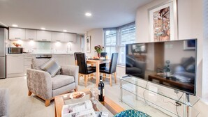A modern living room with a sofa, coffee table, and a TV on a glass stand. There is a dining area with chairs and a table, and a kitchen with cabinets and appliances. - 1 Bed in Whitby (oc-w503) (Whitby)
