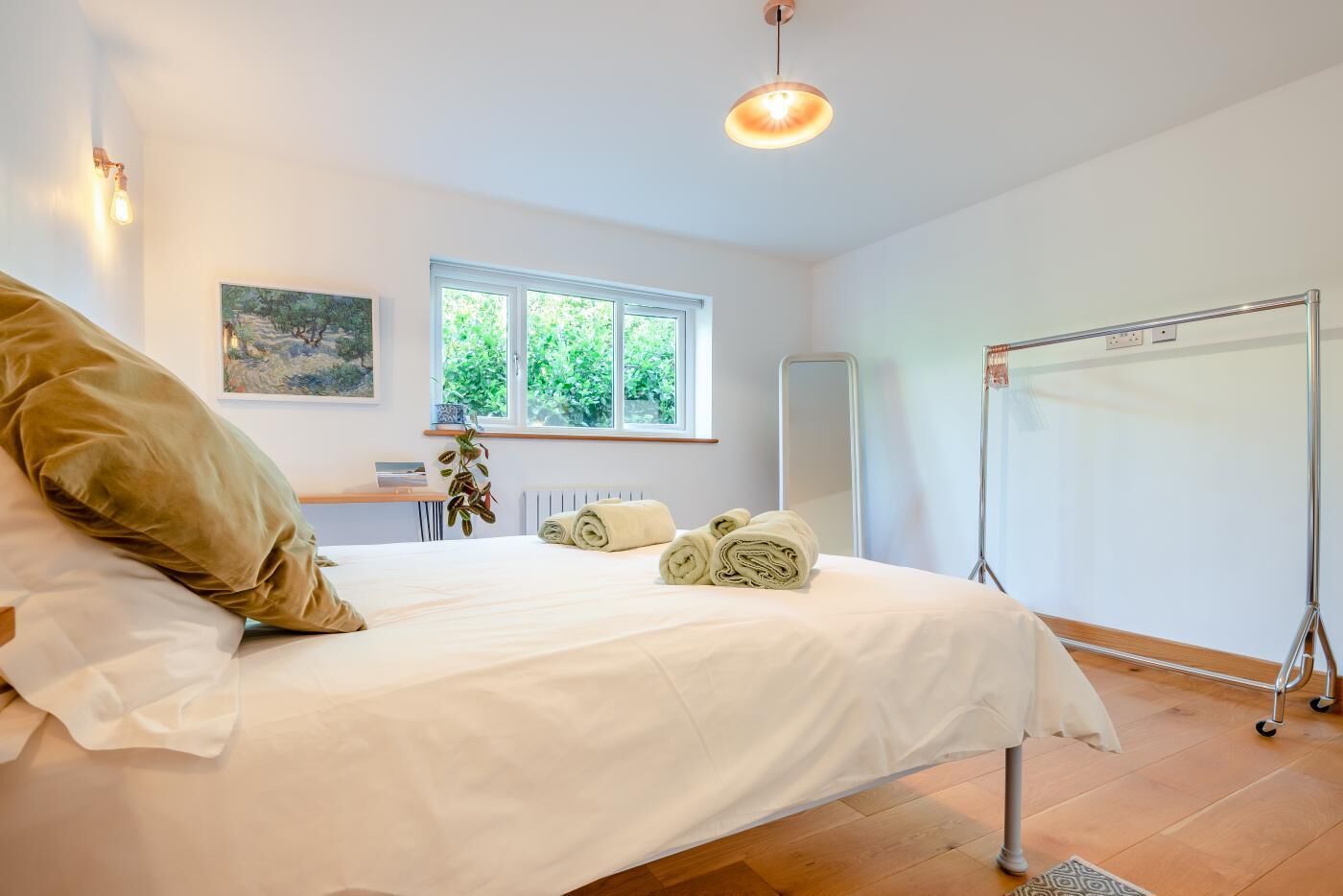 A neatly made bed with rolled towels, a bedside table, a window with a view of greenery, a wall-mounted painting, and a standing mirror.