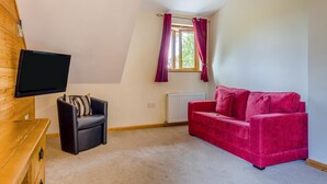 Um quarto com uma poltrona de couro preto, um sofá de veludo vermelho, uma televisão fixada numa parede de madeira, uma janela com cortinas e um radiador.