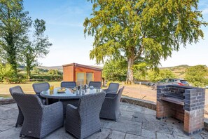 An outdoor dining area with wicker chairs and a glass table, a brick barbecue grill, and a wooden cabin structure. - 9 Bed in Brecon (oc-cvelo) (Crai)