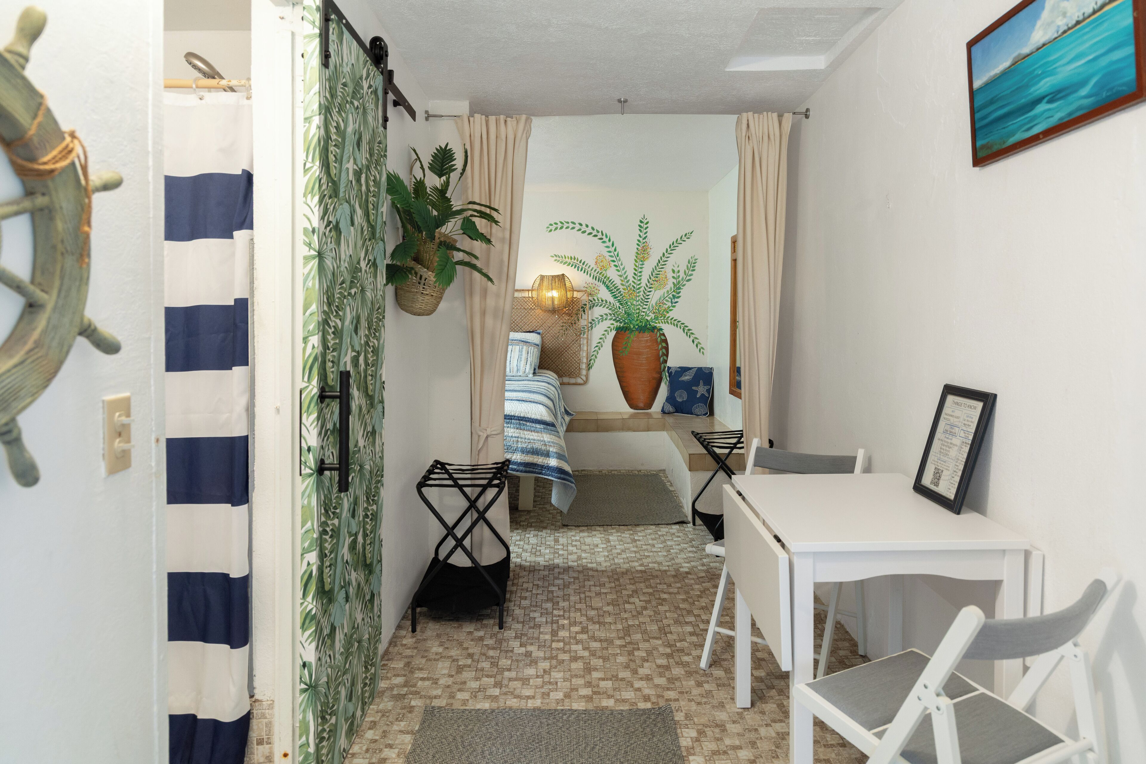 A compact room with a bed, a small table, and chairs. There is a potted plant and a framed picture on the wall.